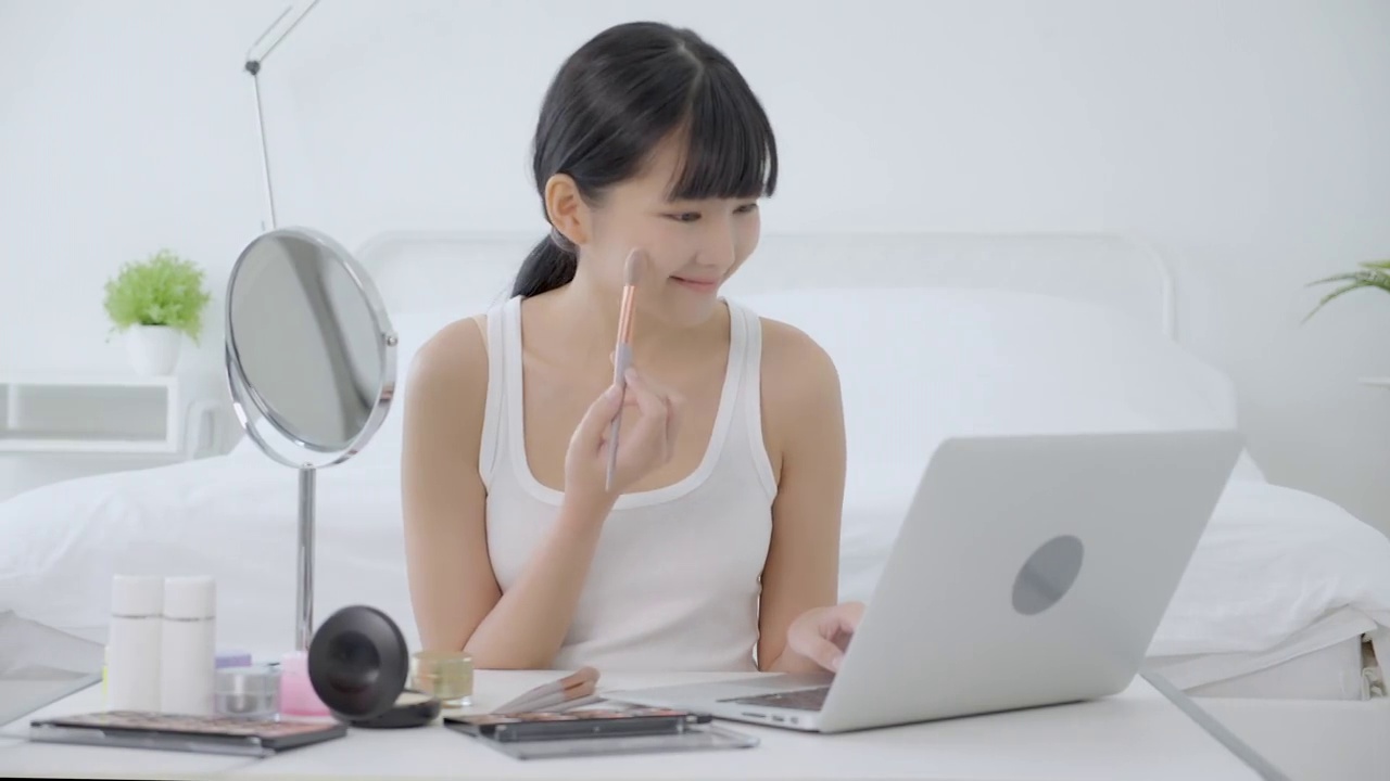 美丽的年轻亚洲女性学习化妆刷在笔记本电脑上与在线教程课程，女性教学和解释化妆与流媒体上的化妆品。视频下载