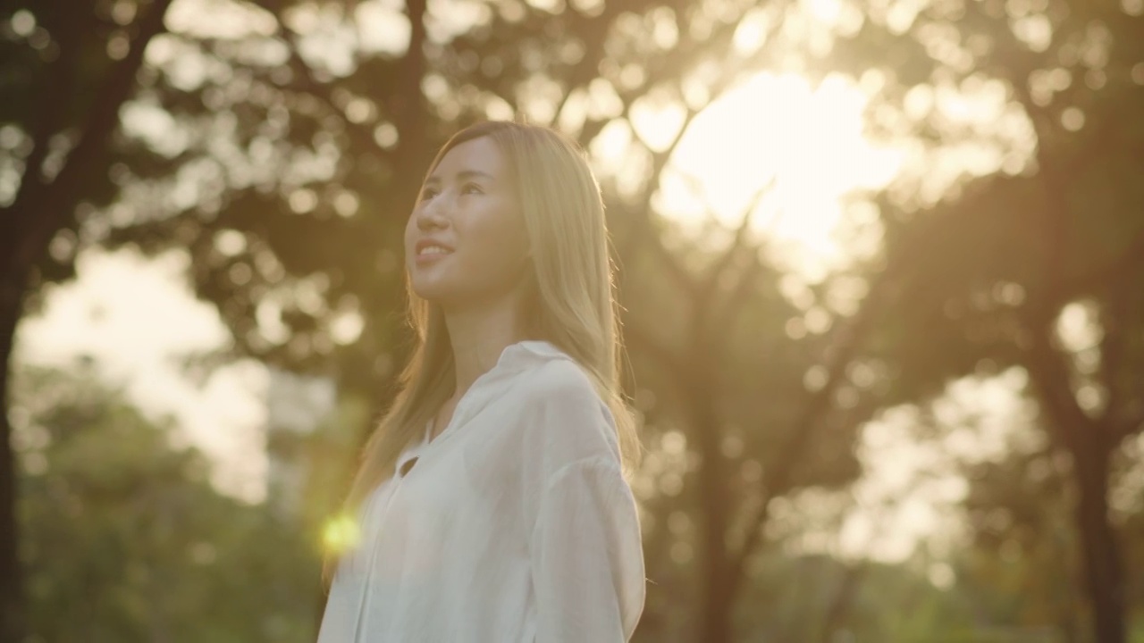 一个美丽的年轻亚洲女性享受微风森林在公园在日落。视频素材