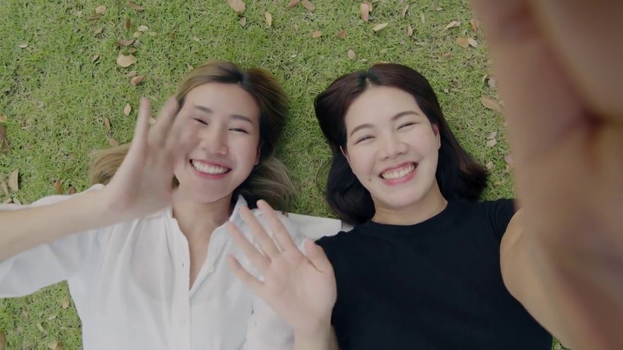 两个年轻的女性朋友一边躺在绿色的草地上享受度假旅行，一边用智能手机自拍视频。视频素材