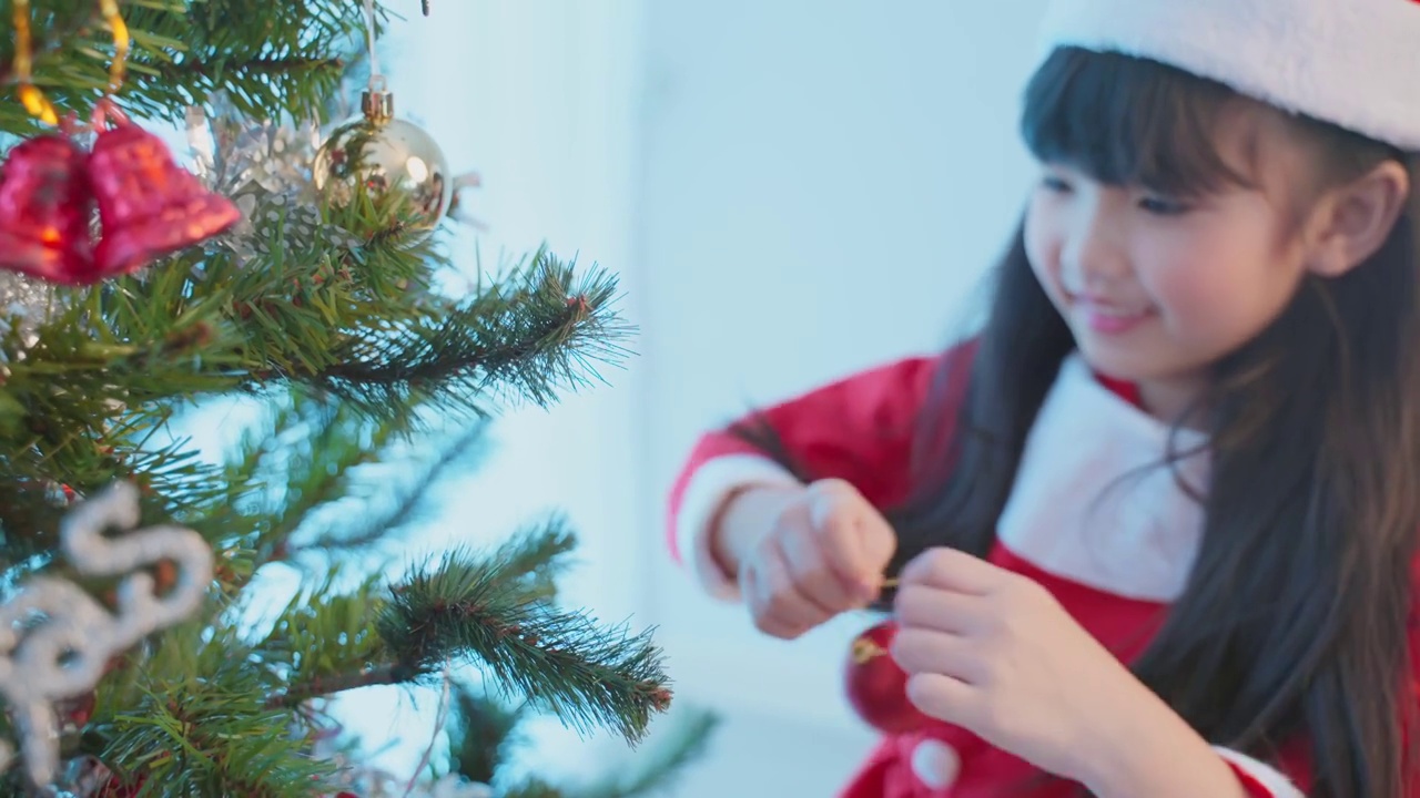 亚洲年轻快乐的女孩孩子戴着圣诞帽，装饰圣诞树。小可爱的孩子穿着红布感到兴奋庆祝节日圣诞节感恩节派对在客厅在房子。视频素材