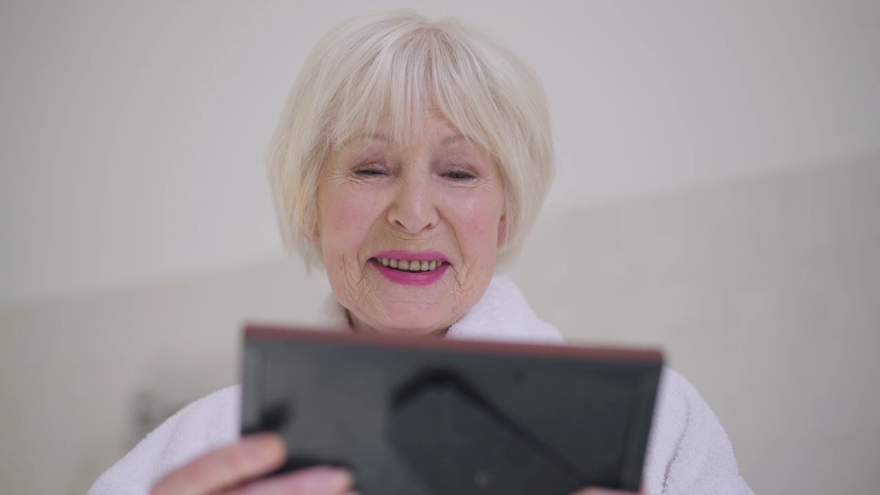 快乐微笑的退休人员看着相框微笑的慢动作的肖像。白种人灰白头发的美丽女人回忆美好的回忆，站在室内，穿着浴袍。视频素材