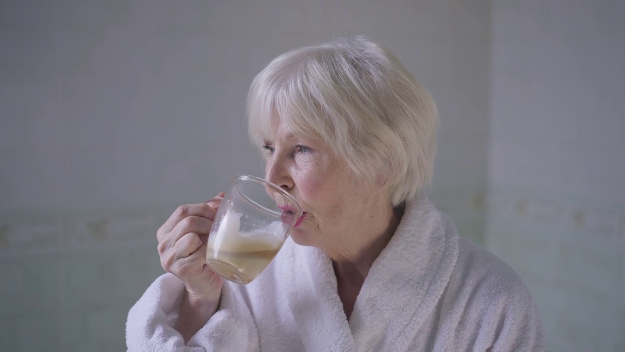 美丽的白发老太太喝着咖啡，慢镜头转过来，微笑着看着镜头。快乐、自信、轻松的白人退休人员在家里的浴室摆姿势与热饮料的肖像。视频素材