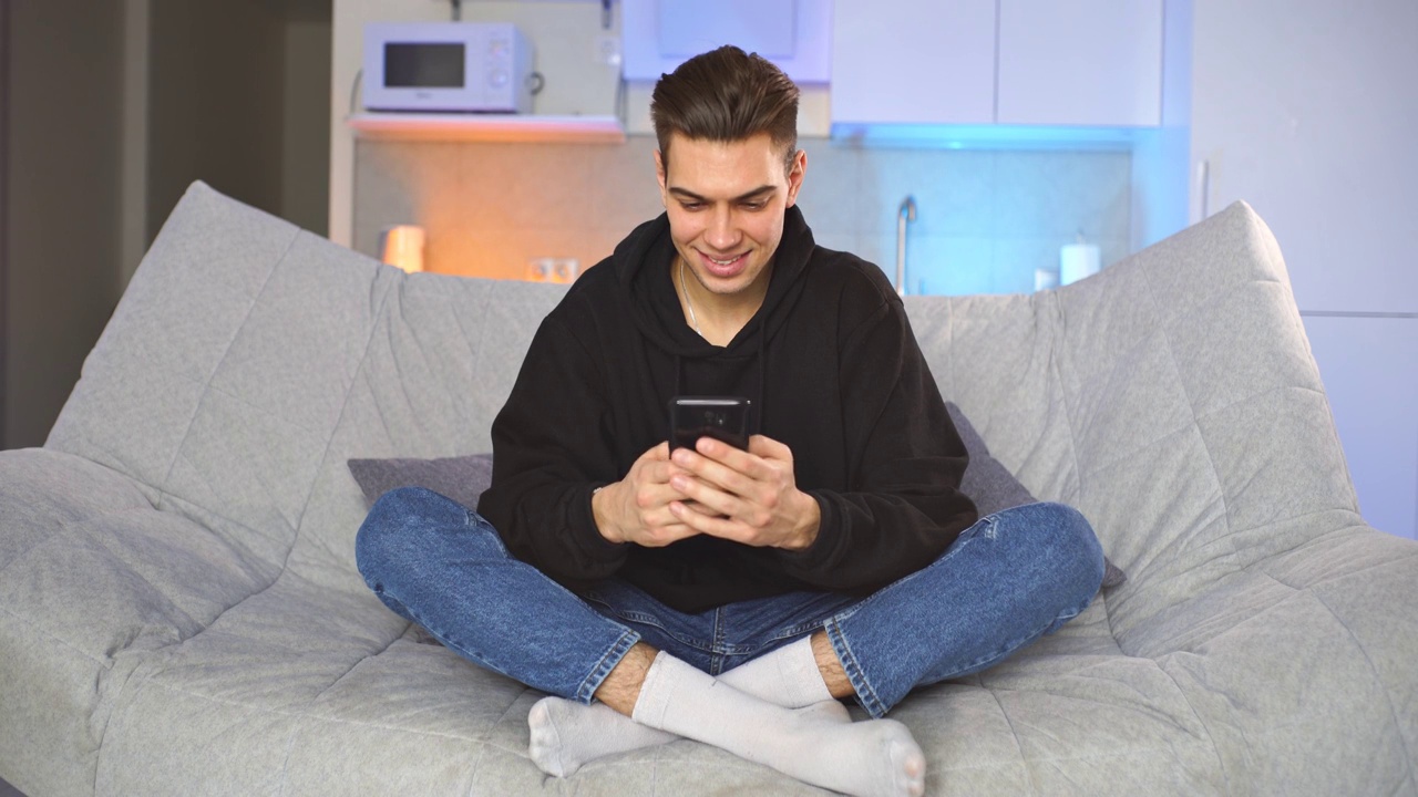 时髦的年轻人坐在家里的沙发上，拿着和使用智能手机视频素材
