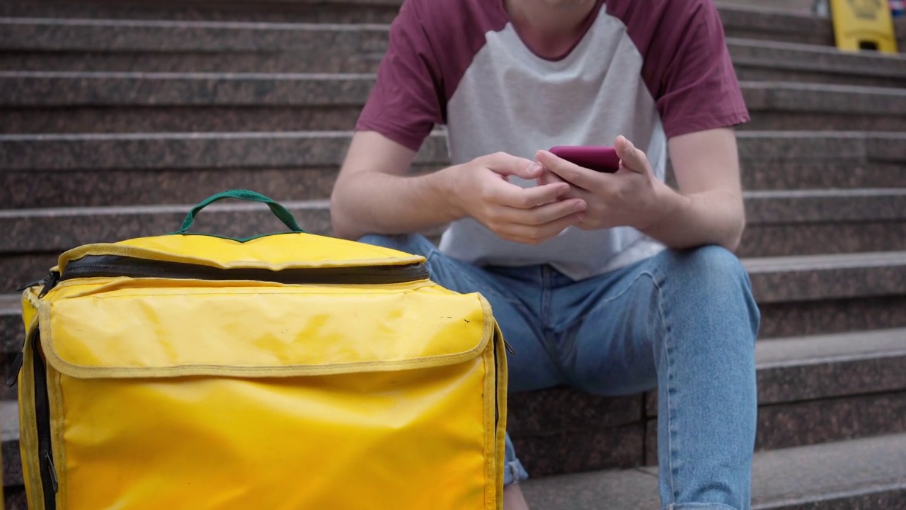 一个面目全非的年轻人坐在楼梯上，背着黄色的送餐背包，用智能手机上网。白人男性快递员在城市户外等待在线订购视频素材