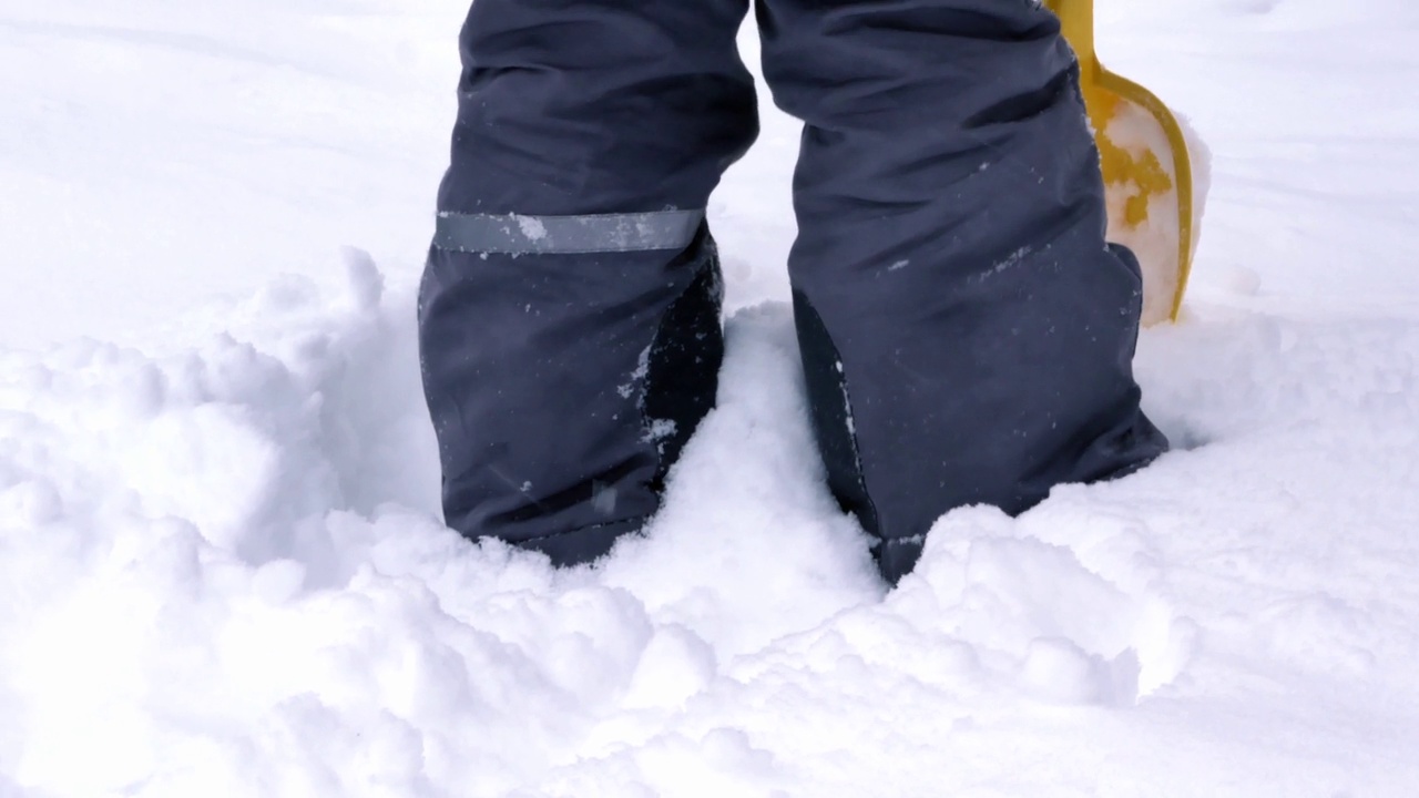 一个孩子的腿上穿着暖和的冬衣，在深深的雪堆中穿行，孩子挥舞着黄色的铲子，下起了雪。快乐的童年，走在清新的空气里视频下载