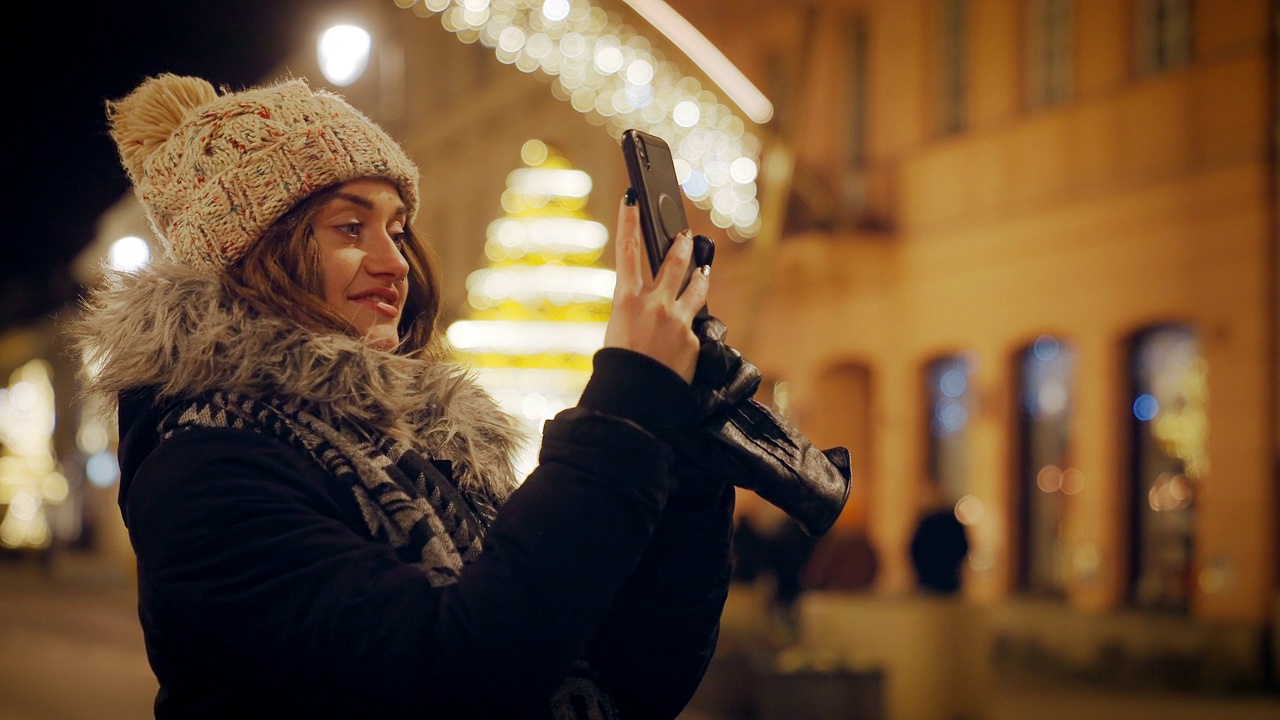 有吸引力的年轻女子，一个游客，享受节日装饰在一个大城市的圣诞节假期。她正在用智能手机自拍，转身拍摄照明照片。视频下载