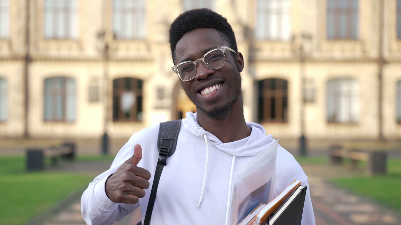 快乐的非洲裔美国学生的肖像，在慢动作拇指向上微笑着看着镜头。戴眼镜、自信、聪明、帅气的男子在大学校园摆姿势，露出露齿的微笑。视频素材