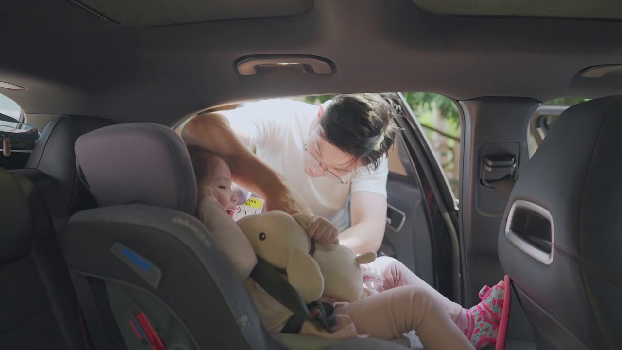 蹒跚学步的小女孩坐在汽车安全座椅上，父亲把她扣在汽车安全座椅上视频素材