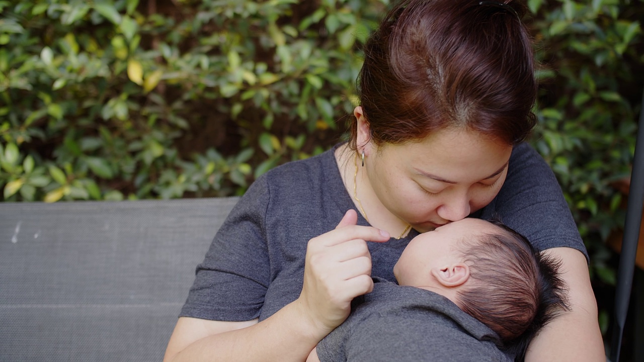 幸福年轻的亚洲母亲抱着亲吻她漂亮的新生婴儿婴儿女孩躺在家里后院的秋千上，孩子，父母，童年，生活，母亲，母亲的概念视频下载