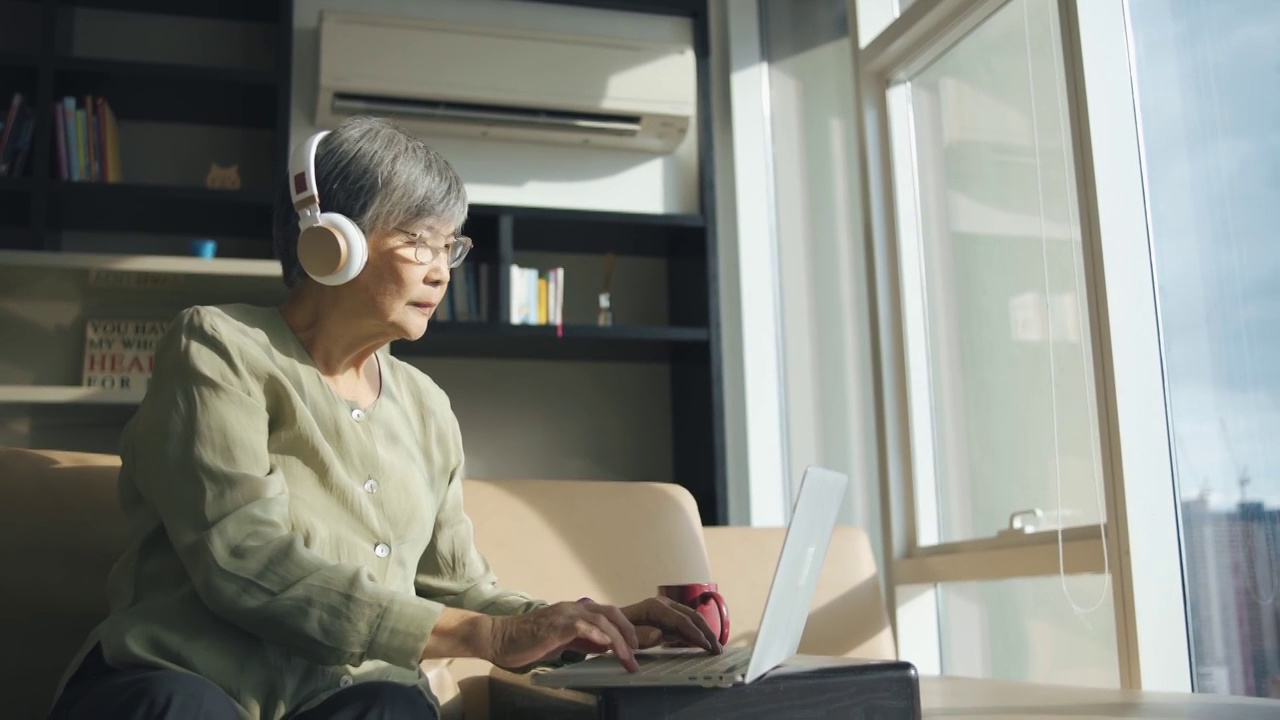 亚裔中国高级女性在她城市公寓的客厅里用耳机使用笔记本电脑进行在线学习视频下载