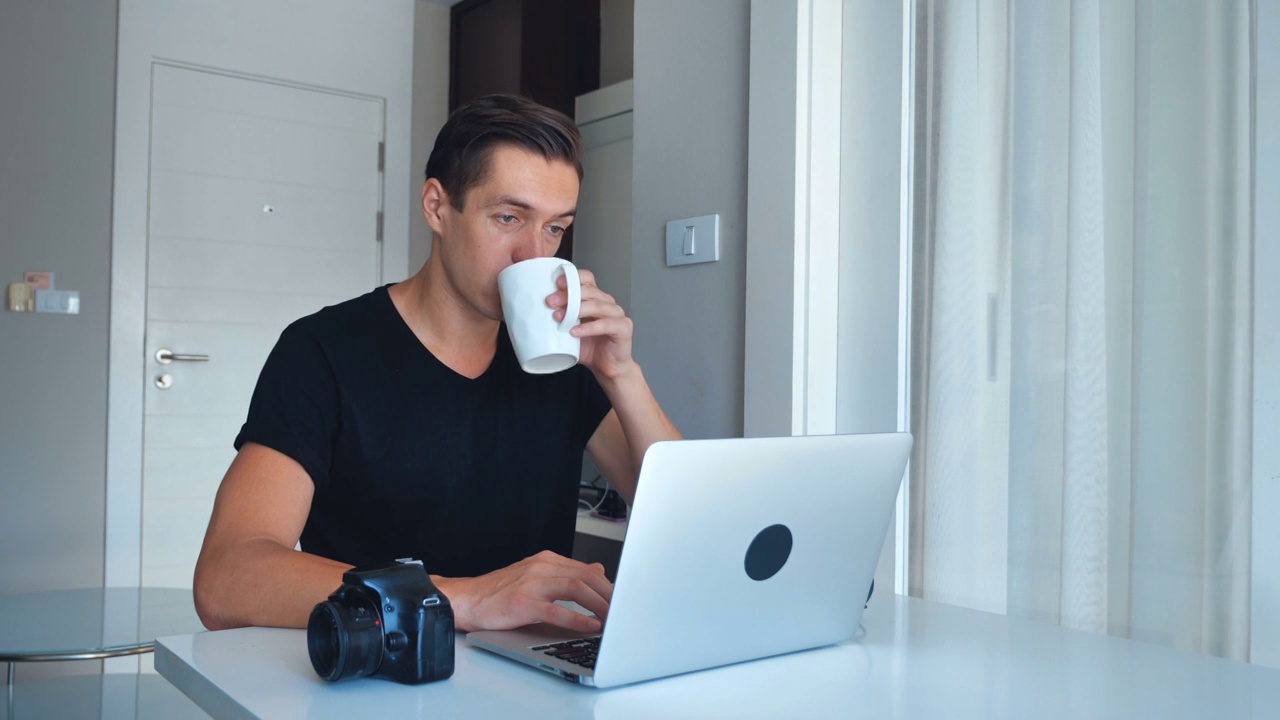 年轻的摄影师在家里喝着咖啡，用笔记本电脑处理图像。相机，外置硬盘在旁边。视频素材