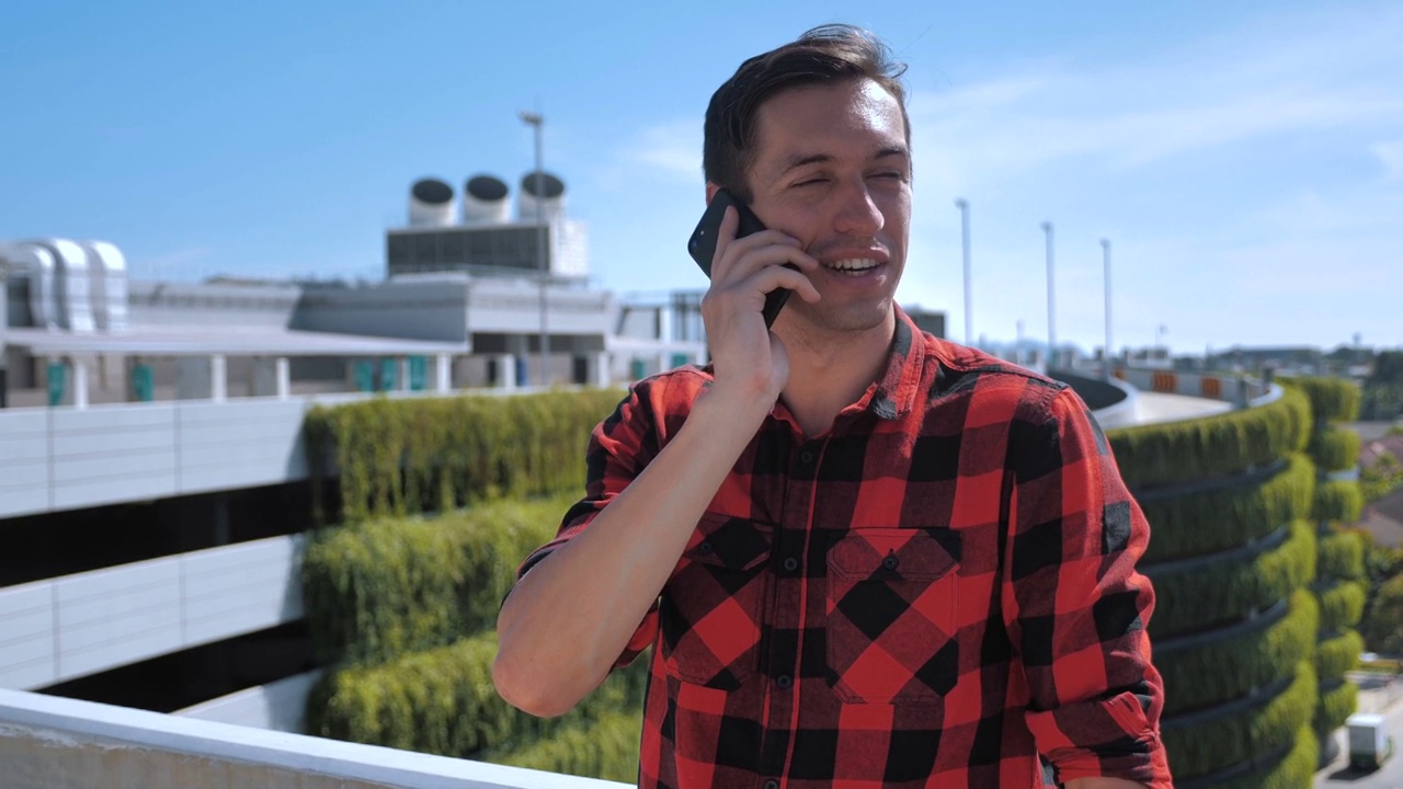 身着格子衬衫，面带微笑的年轻男子站在一座建筑的屋顶上，在城市的背景下打电话视频素材