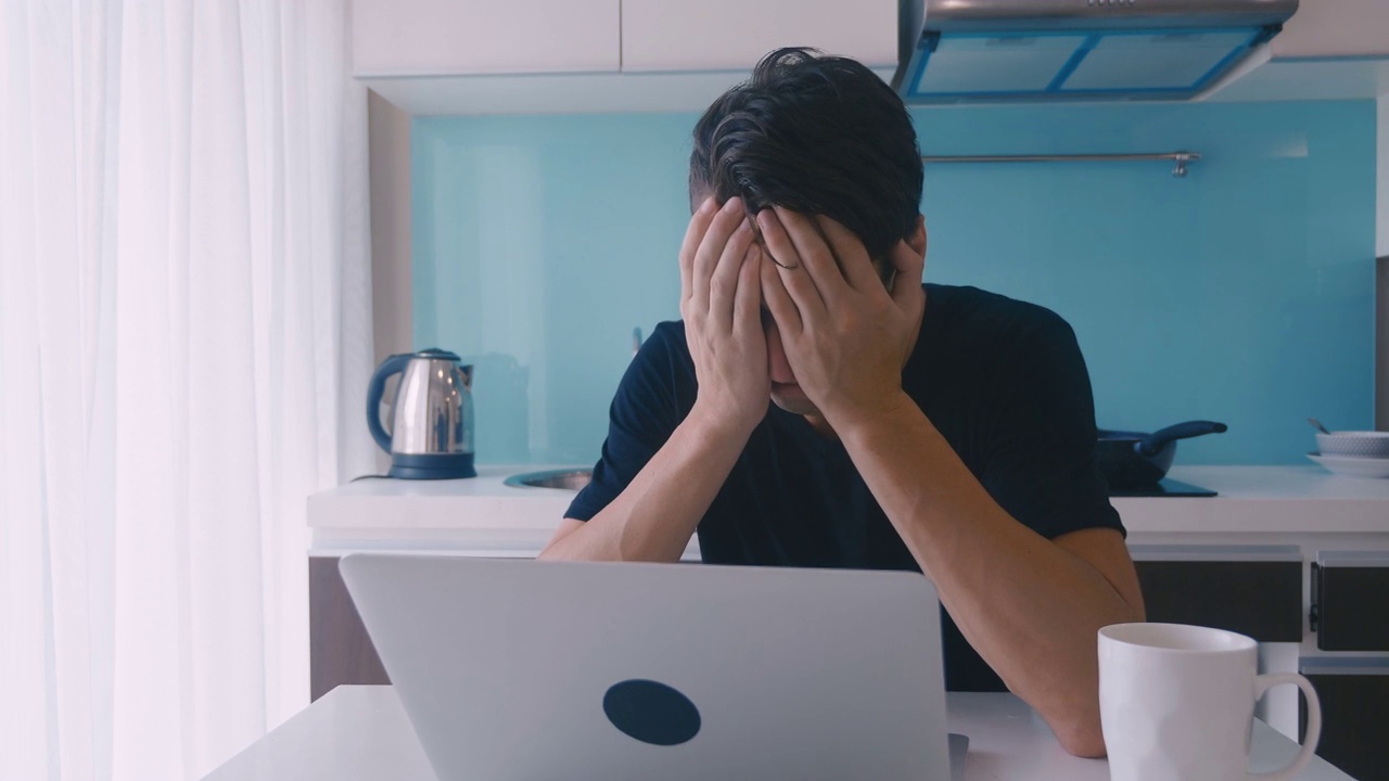 抑郁的年轻人在家里厨房里用笔记本电脑工作。一个男人头痛。视频素材