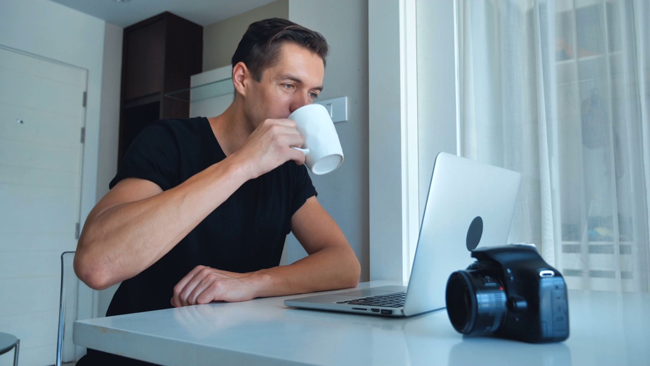 年轻的摄影师在家里喝着咖啡，用笔记本电脑处理图像。相机，外置硬盘在旁边。视频素材