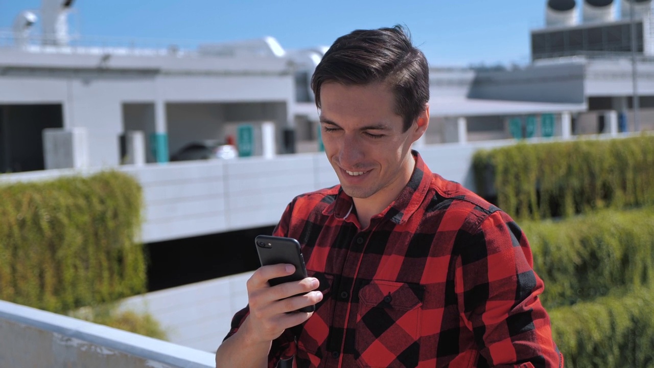 年轻微笑的男子在格子衬衫在电话短信现代智能手机在屋顶在城市背景视频素材