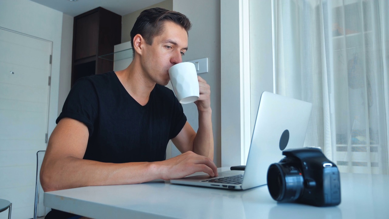 年轻的男摄影师在家里喝着咖啡，用笔记本电脑处理图像。视频素材