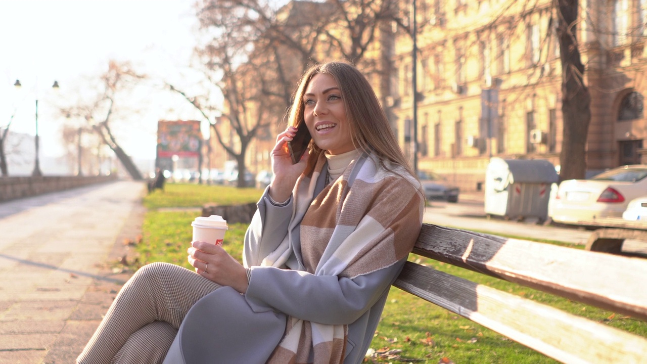 微笑的年轻女子喝着外卖咖啡，在城市公园里用智能手机聊天视频下载