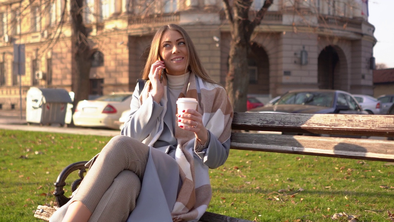 微笑的年轻女子喝着外卖咖啡，在城市公园里用智能手机聊天视频下载
