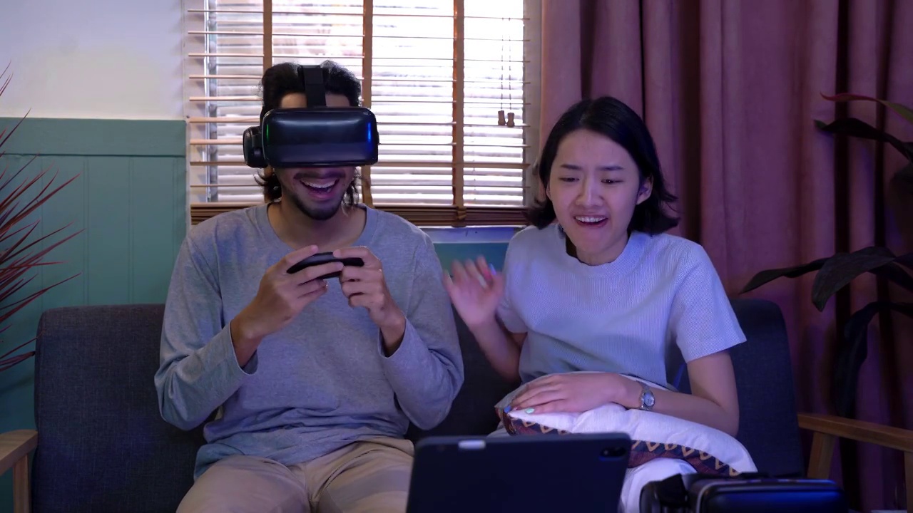 亚洲男子在客厅里戴着虚拟现实眼镜。戴着耳机观看的人们感到高兴。内部与照明从笔记本电脑屏幕。男子用手柄玩游戏。视频下载