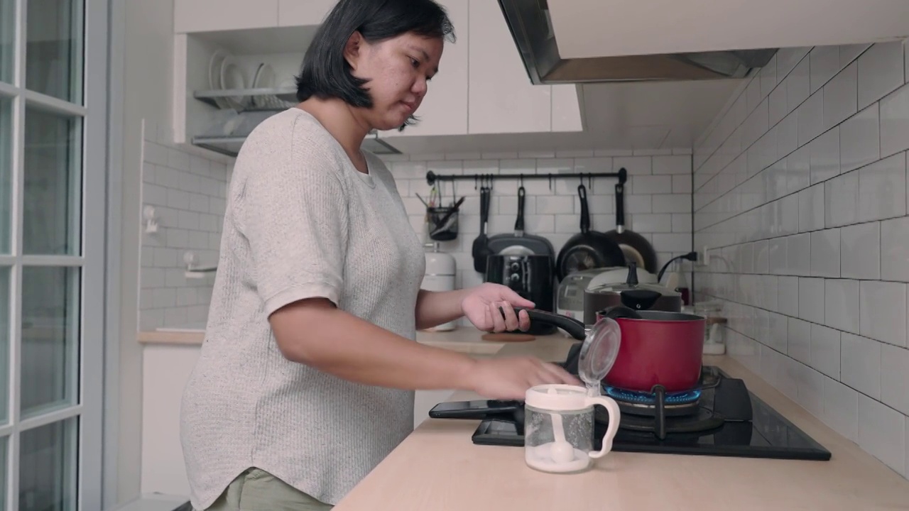 亚洲妇女在厨房做饭，同时看手机的食谱视频素材