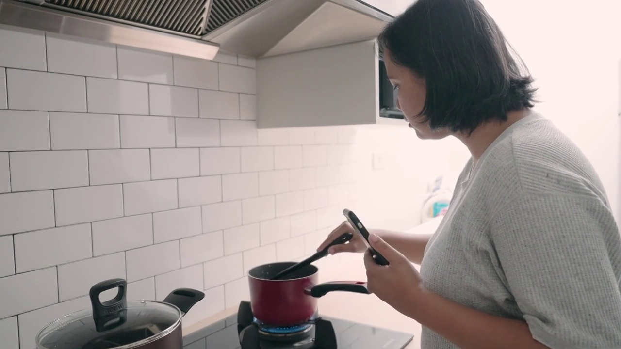 亚洲妇女在厨房做饭，同时看手机的食谱视频素材