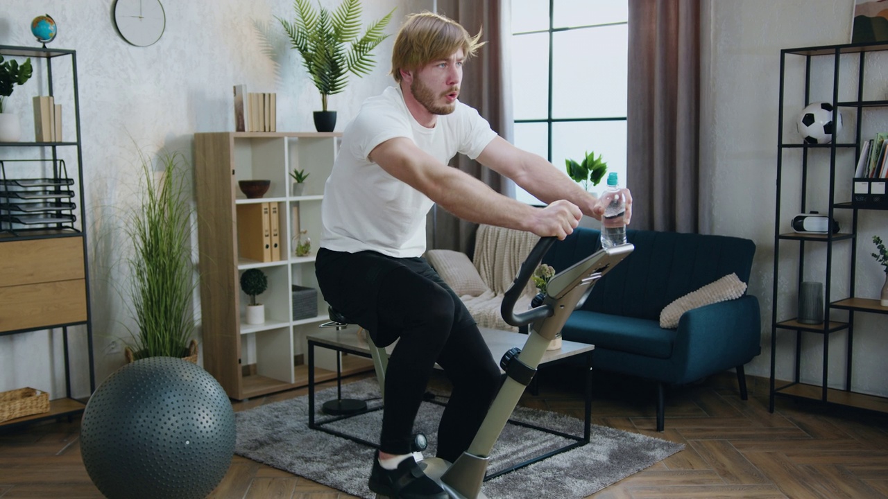 长得好看、精力充沛、肌肉结实、留着大胡子的家伙穿着运动服，在早上的家庭锻炼中骑着固定自行车视频下载
