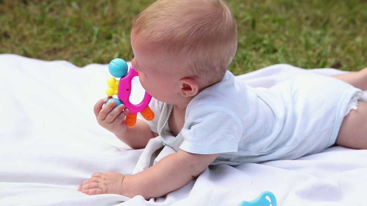 初生女婴躺在毯子上嚼拨浪鼓，夏日户外。婴儿在户外玩耍。婴儿，婴儿，孩子休息，玩耍，学习爬行。母性、牙齿、婴儿概念视频下载
