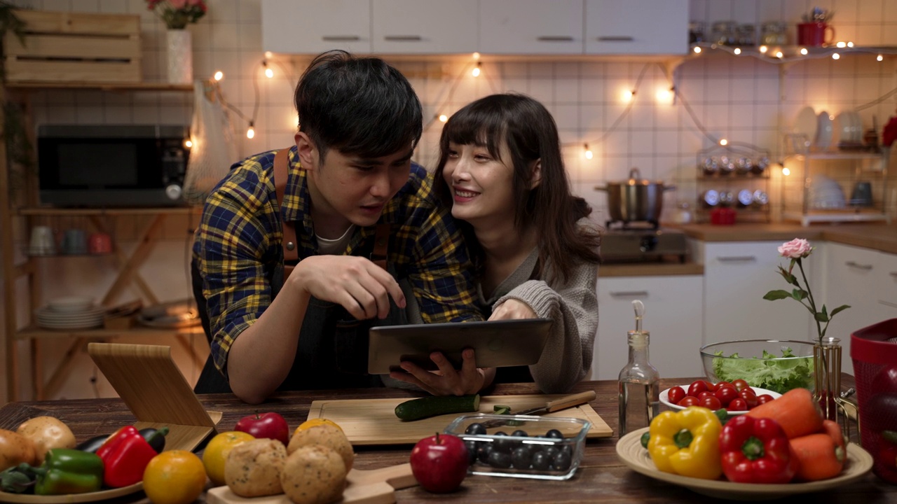在家里的厨房里，一对快乐的亚洲情侣用一个触摸板一起计划和决定情人节晚餐的食谱视频下载