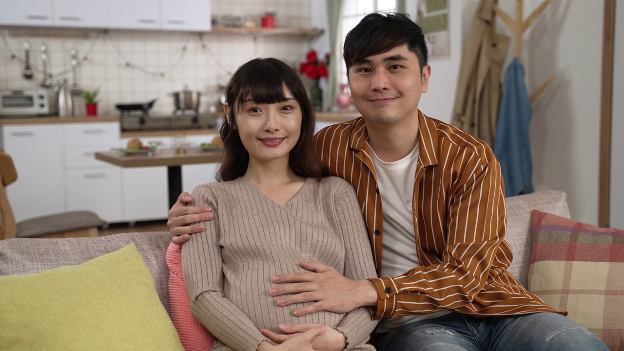 年轻微笑的亚洲夫妇的肖像看着相机在一个舒适的客厅在家里。丈夫把手放在怀孕的妻子肚子上。视频素材
