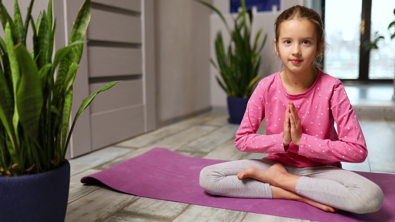 可爱的小女孩坐在健身垫上，合十礼摆莲花姿势，在家里做瑜伽视频素材