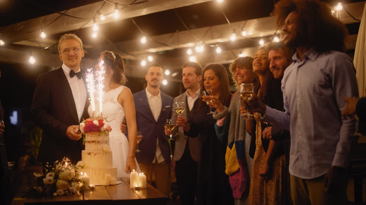美丽的新娘和新郎与多民族的朋友在晚会上庆祝婚礼。新婚夫妇为婚姻幸福祝酒，站在餐桌前，桌上放着蛋糕和烟花。视频素材