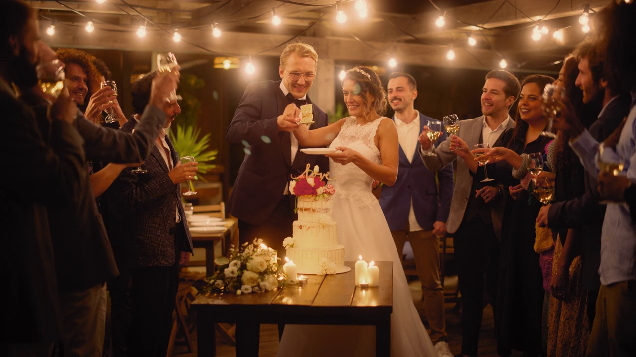 美丽的新娘和新郎与多民族的朋友在晚会上庆祝婚礼。已婚夫妇站在餐桌前，亲吻和切婚礼蛋糕。视频素材