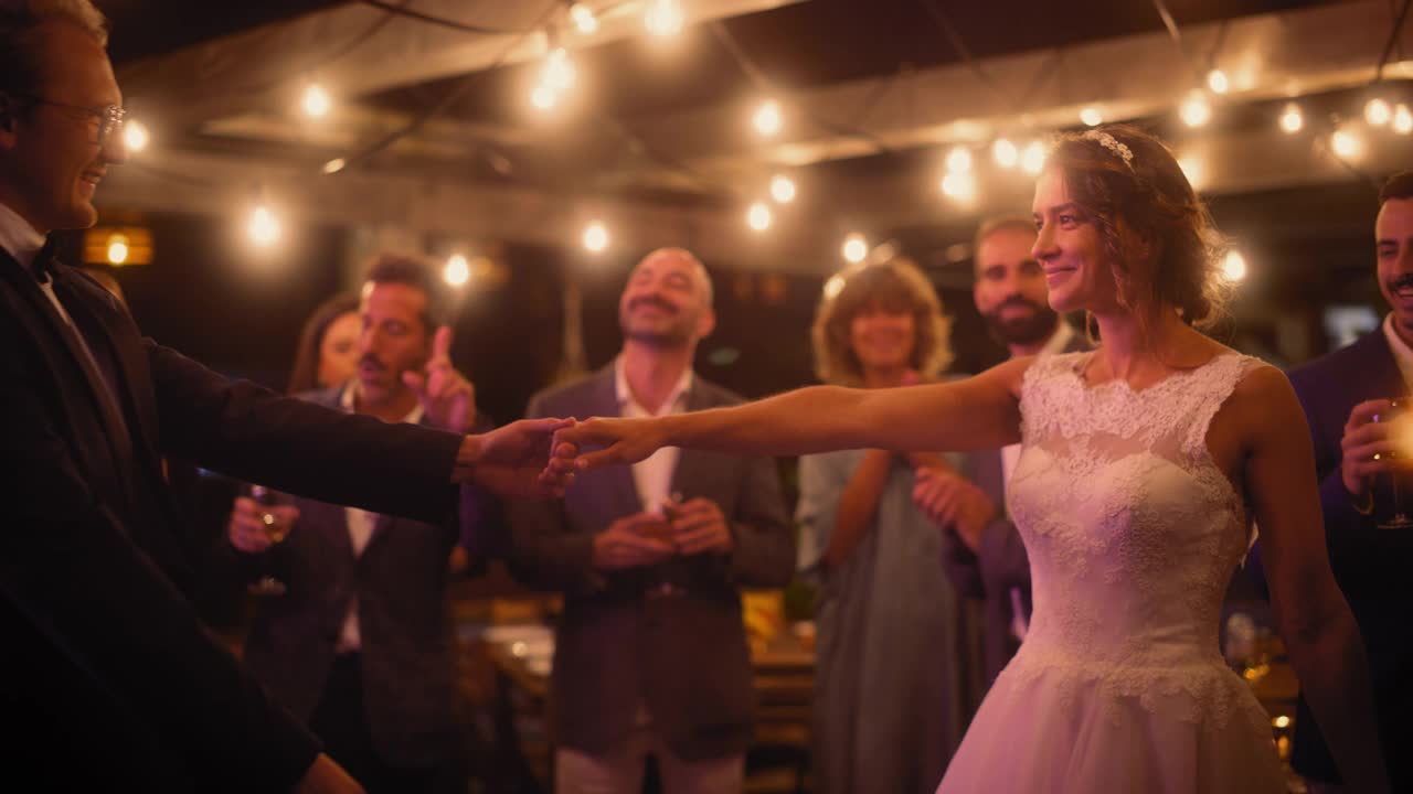 美丽的新娘在白色礼服和新郎在时尚的黑色套装庆祝婚礼在一个晚上的招待会上。新婚夫妇和最好的多种族朋友在一个场所跳舞。视频下载