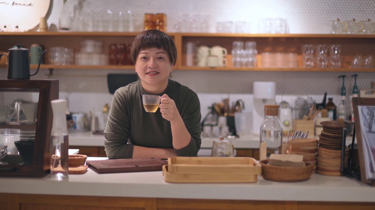 亚裔华人小企业主拿着茶杯站在厨房柜台前微笑着看着镜头视频下载