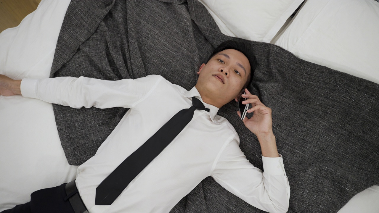 俯视图不知所措的亚洲商人躺在床上，拿着电话听着，无精打采地在家里的卧室里回复视频素材