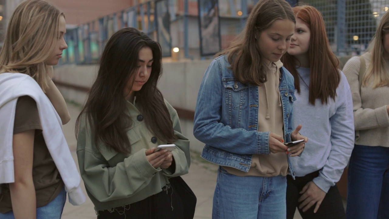 五个放松的十几岁女孩站在街上，看着自己的手机视频下载