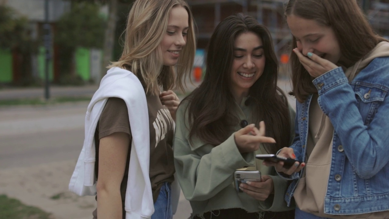三个放松的十几岁女孩站在街上，看着自己的手机视频下载