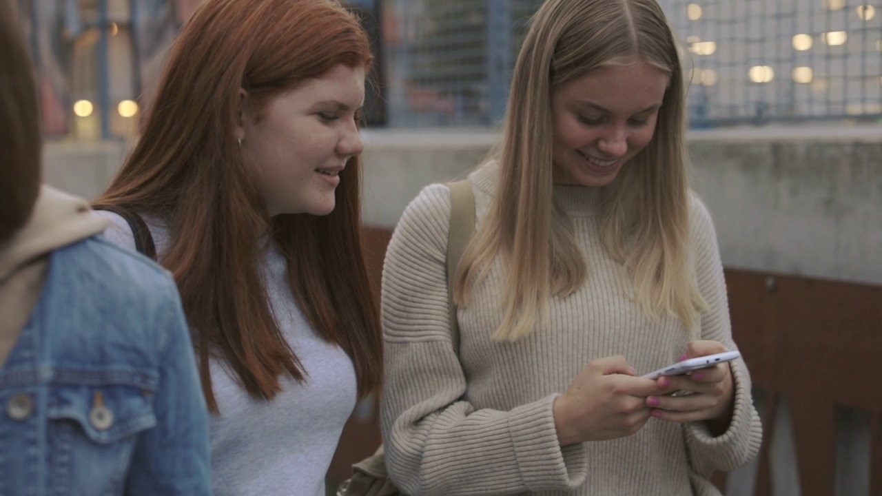 两个放松的十几岁女孩站在街上，看着自己的手机视频下载