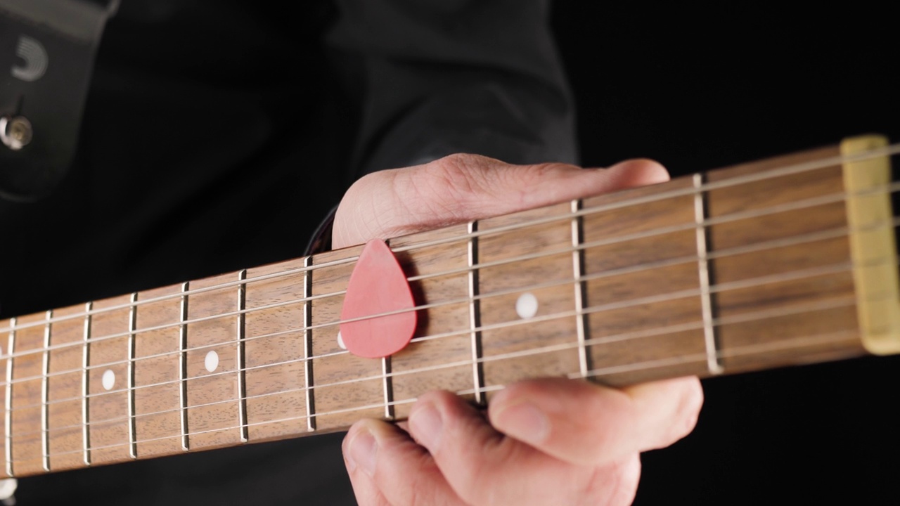 吉他手在黑色的背景下，从浅棕色的六弦电吉他的弦上拿起一个红色的拨片。在家做点音乐的概念。声音艺术的自由时间。中间。4 k视频素材