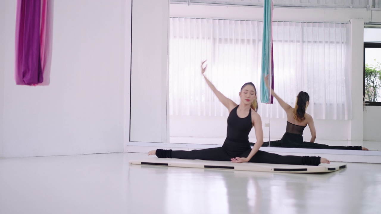 年轻的亚洲妇女在瑜伽垫上伸展双腿，在白色的房间周围有镜子。练习瑜伽可以增强柔韧性和伸展肌肉，锻炼身体，保持健康，穿着运动服，裤子和上衣。视频素材