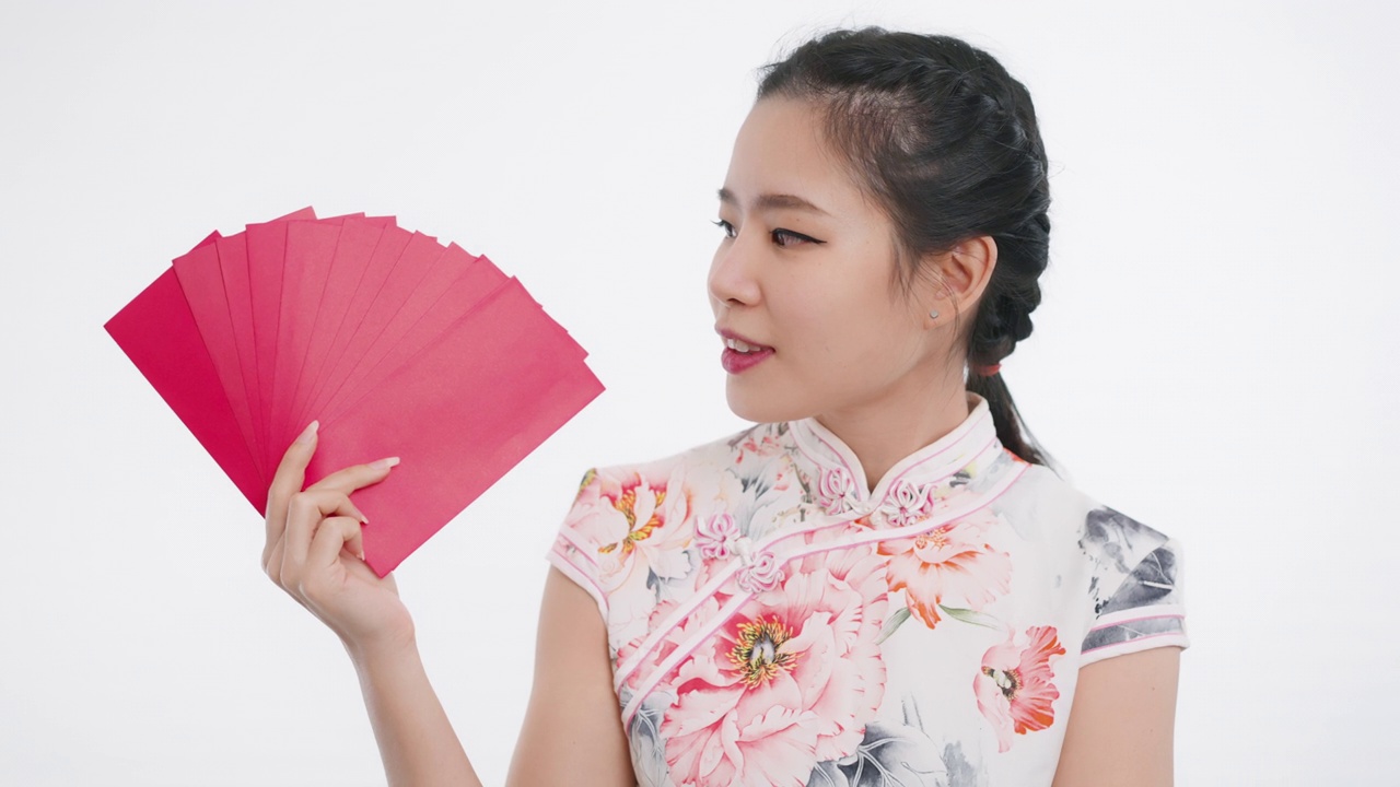 中国/亚洲美女展示新年快乐，富有，幸福，和幸运的红包，她站在白色背景，穿着旗袍白色花卉图案的服装手视频下载