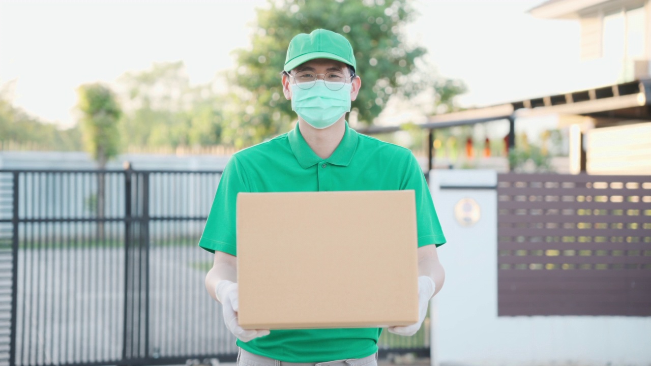 冠状病毒概念，帅气的亚洲送货男子穿着绿色制服，拿着包裹纸板箱，戴着防护口罩和医用橡胶手套在客户房前。4 k的决议视频下载