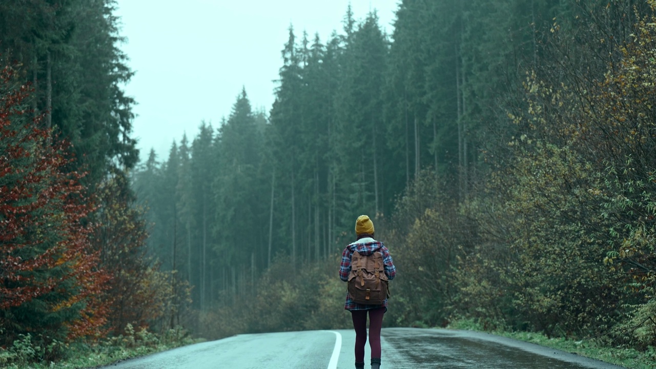 背景:在寒冷潮湿多雾的秋日里，一个背双肩包的女人走在户外的路上，等着一辆车。旅游旅行搭山旅游自然度假旅游自由旅游视频素材
