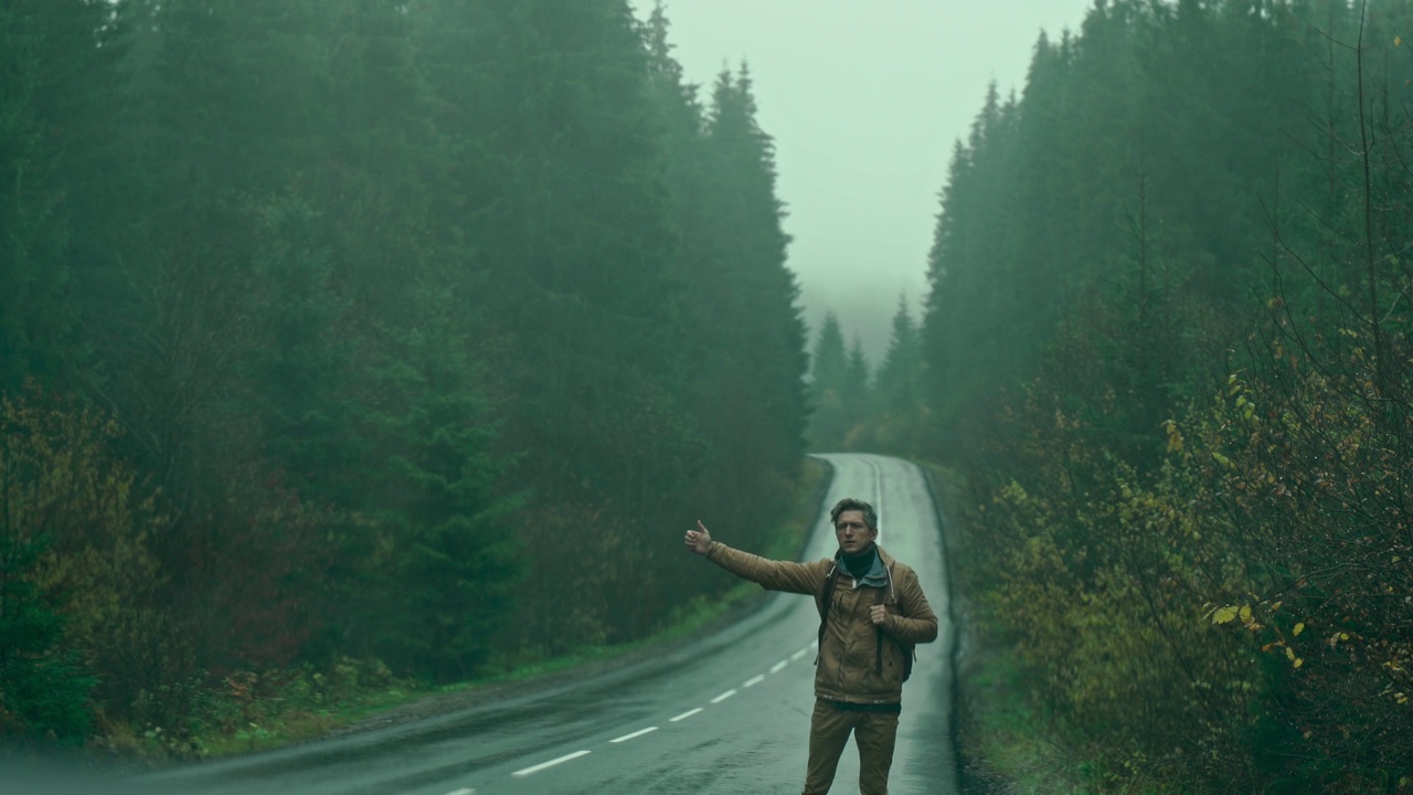 在寒冷潮湿多雾的秋日，一名背包旅行者站在森林的山路上，搭上了一辆汽车。人向前看，等待视频素材