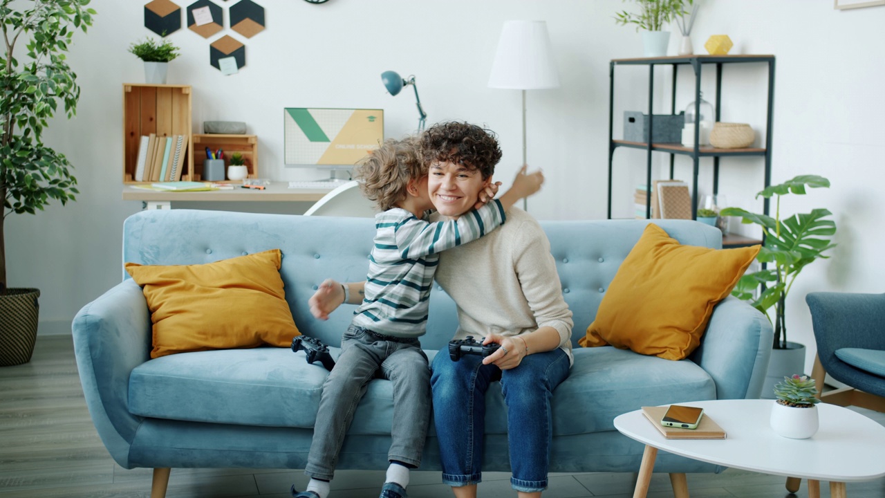 可爱的男孩玩电子游戏的肖像与母亲赢得拥抱父母在家里室内视频素材