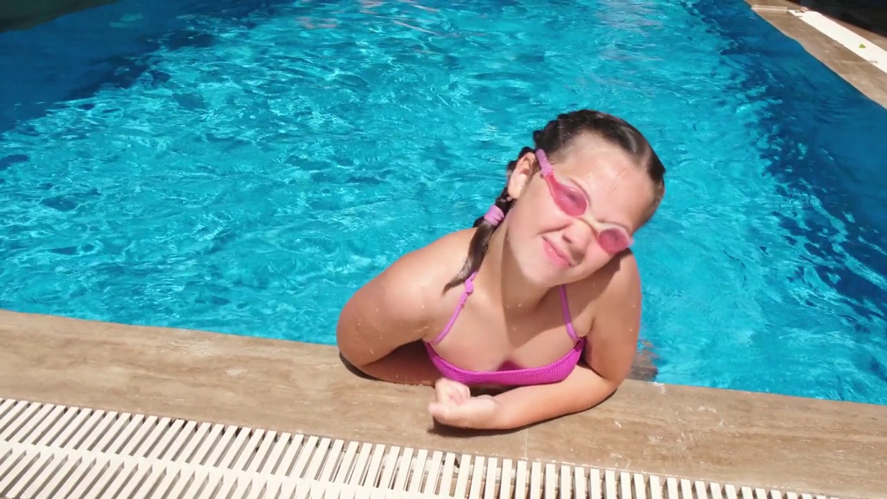 微笑的少女在游泳池的水杯里。女孩坐在池边做鬼脸。炎热阳光的度假概念。慢动作视频下载