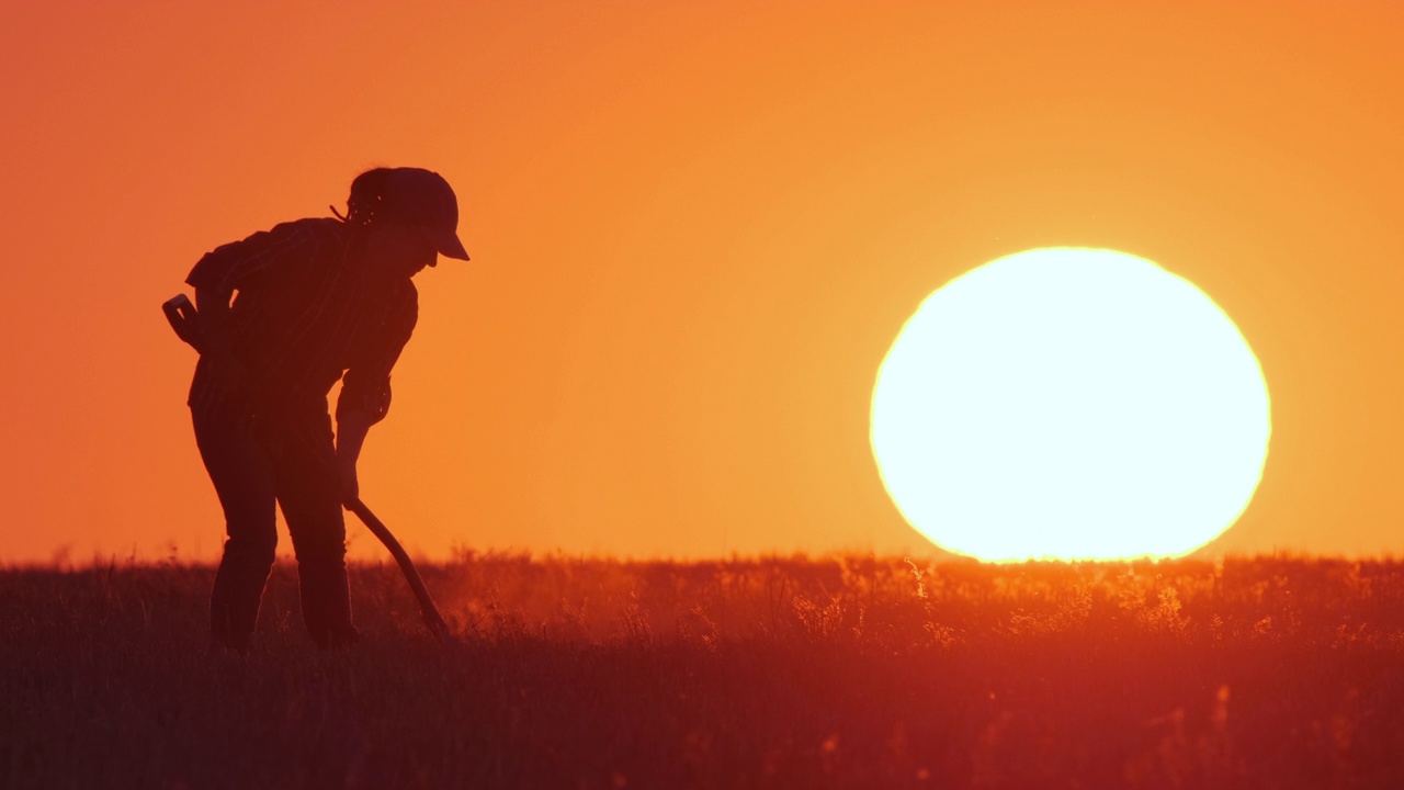 穿着橡胶靴的农民或园丁的剪影，在橘黄色的日落天空和大太阳的背景下，用铲子挖掘地面视频下载