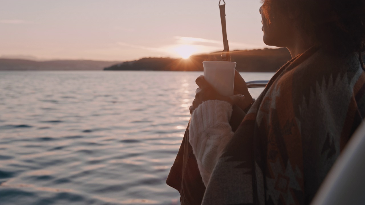 日落时，一名年轻女子裹着一条毯子，在帆船的甲板上喝茶视频素材