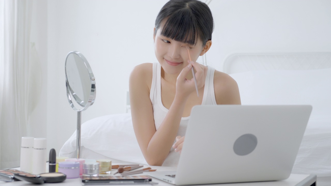年轻的亚洲美女与学习化妆刷眼影笔记本电脑与教程课程在线，女性教学和解释眼影与化妆品在流媒体上化妆。视频下载