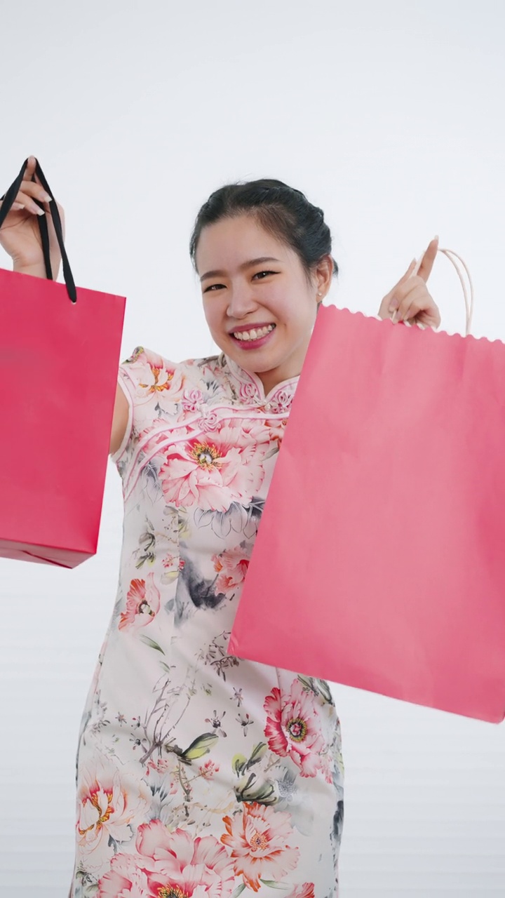 中国/亚洲妇女在中国新年期间，双手拿着红色和粉红色的购物袋，白色背景，穿着旗袍白色花卉图案连衣裙视频下载